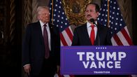 Two men in suits stand by a podium that has a "Trump Vance" sign on it.