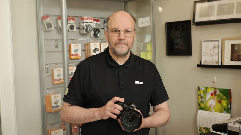 En man i svart pikétröja med Nikon-logga håller en kamera i en butik med kameratillbehör och fotoprodukter.