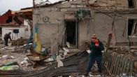 Woman in green pants and top holding a smartphone walking in front of the remains of a building damaged in an air strike.