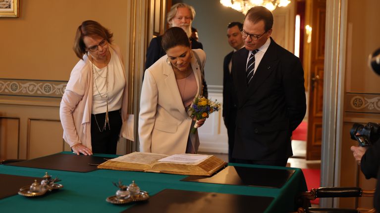Kronprinsessan Victoria, prins Daniel och två andra personer står runt ett uppslaget dokument på ett bord i ett inrett rum.