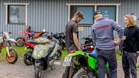 Tre personer står intill flera motorcyklar och mopeder utanför en byggnad med grå panel och skyltar på fönstren.