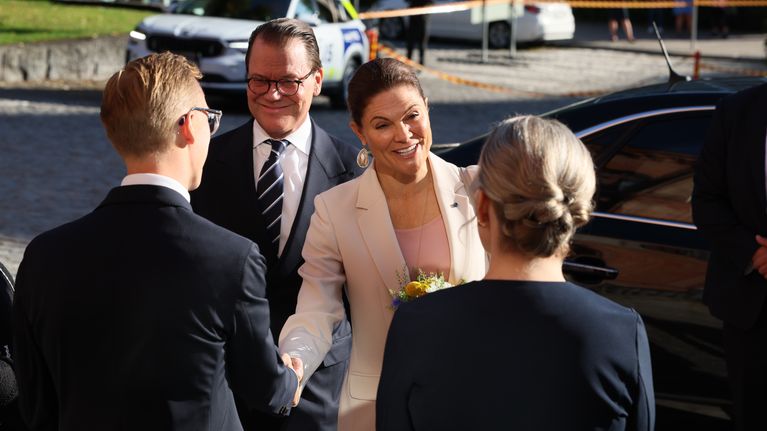 Kronprinsessa Victoria i ljus kavaj skakar hand med en person utomhus, omgiven av flera personer och parkerade bilar.