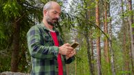 En man står i skogen och tittar på sin mobiltelefon. Han bär en grön- och svart-rutig skjorta över en röd tröja. Bakgrunden består av träd.