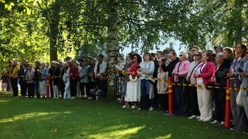 En grupp människor står bakom ett rödgult avspärrningsband utomhus och tittar åt samma håll. Träd och gräs syns i bakgrunden.