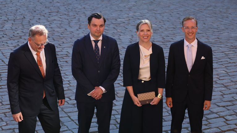 Fyra personer står uppställda på en stenbelagd yta, samtliga klädda i kostym eller kavaj.