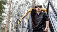 En ung pojke (Emil Pääkkölä), fotograferad nerifrån, står i dörren av en skogsmaskin, en skördare, med den snöiga skogen i bakgrunden.