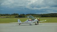 Two people stand by a small white airplane parked on a runway with a glass hatch open and a pilot seated inside.