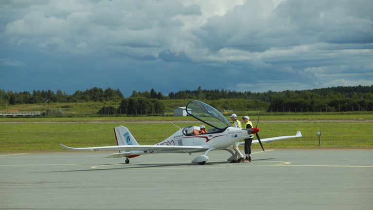 Two people stand by a small white airplane parked on a runway with a glass hatch open and a pilot seated inside.