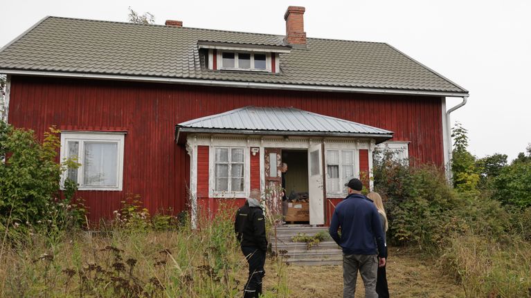 Tre personer står framför ett röd- och vitmålat hus med öppen dörr, omgivna av hög växtlighet.