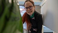 Pitkähiuksinen, silmälasipäinen naisdiakoni katsoo sisään lokerikkoon, kuva otettu lokerikon sisältä ulospäin, etualalla täysi muovikassi.