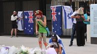 Aitaan on kiinnitetty Australian ja Israelin lippuja. Kaksi ihmistä on kumartunut maassa olevien kukkien puoleen. Taustalla kaksi ihmistä halaa. 
