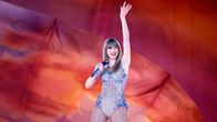 Taylor Swift i en glittrig bodysuit står på en scen och vinkar. Hon ler och håller i en mikrofon.