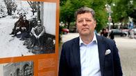 En man i kostym står utomhus bredvid en informationsskylt med historiska fotografier.