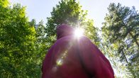 Lastensuojelulaitoksen entinen työntekijä metsässä, aurinko paistaa kirkkaasti puiden lomasta.