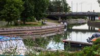 Båtar ligger förtöjda vid en kanal med en bro i bakgrunden, omgiven av träd och grönska.