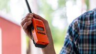 Henkilö pitää oranssia radiopuhelinta kädessään ulkona, taustalla näkyy punainen rakennus ja vihreää puustoa.