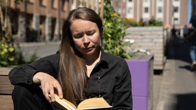 Nainen istuu penkillä ja lukee kirjaa. Penkin vieressä istutuksia. 