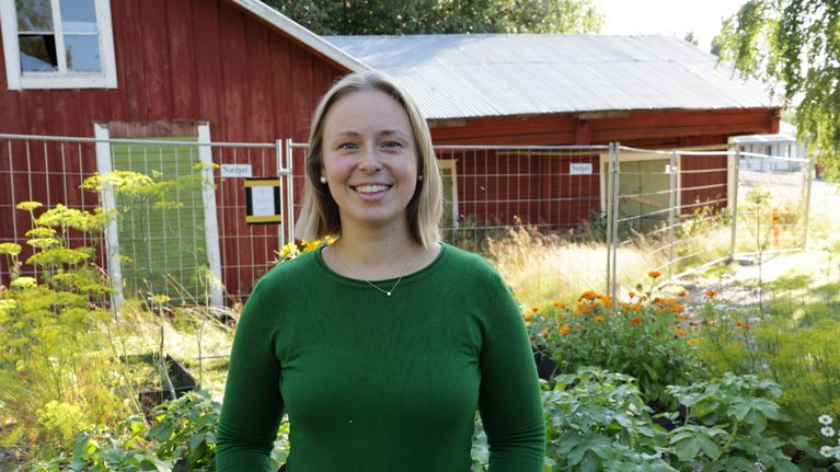 En leende kvinna i grön tröja står framför grönskande odlingslådor.