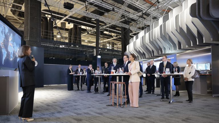 En grupp personer står och lyssnar på en talare i en modern inomhuslokal.