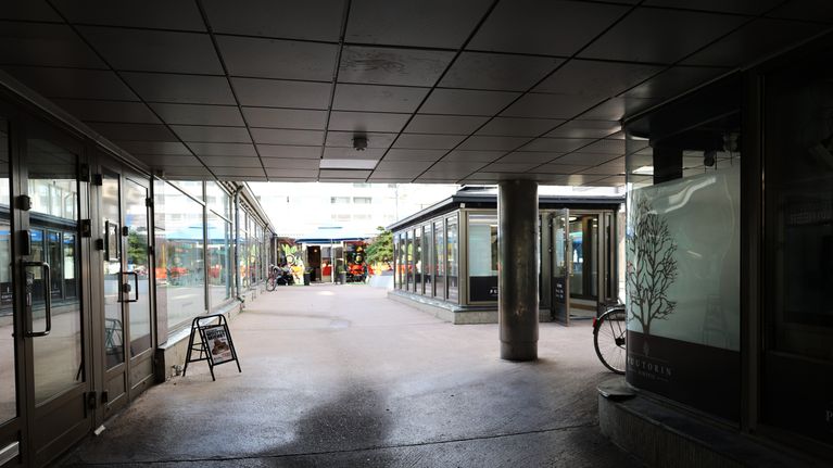 Gård mellan två byggnader med stora fönster, uteservering med blått parasoll och ett flerbostadshus i bakgrunden.