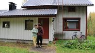 Colegeasuun pukeutuneet tummahiuksiset mies ja nainen seisovat vanhan, valkoisen rintamamiestalon edessä ja nojaavat toisiinsa.