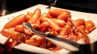 Valkoisella tarjoiluvadilla on useita pieniä nakkeja ja metalliset ottimet.