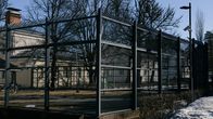Kuvassa on ulkona sijaitseva, korkean metalliaidan ympäröimä pieni liikunta- tai pelialue. Taustalla näkyy puita ja rakennuksia, joista yksi on savupiipulla varustettu. 