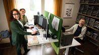 Ett kontor där två personer står vid sina skrivbord och en sitter ned. Alla arbetar vid datorer.