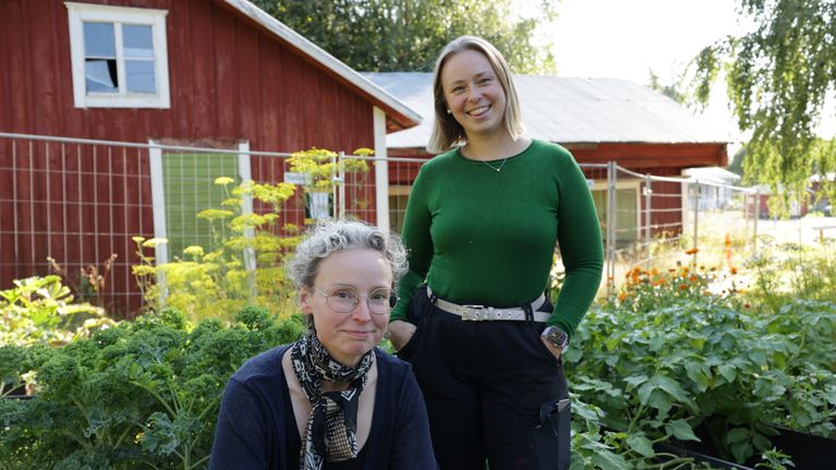 Två leende kvinnor framför grönskande odlingslådor och rödmålade byggnader.