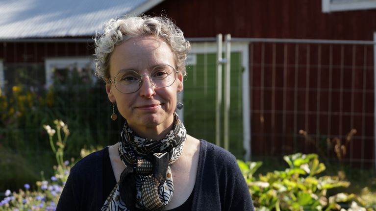 En småleende kvinna med grått lockigt hår fotograferad utomhus i kvällssol.
