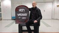 Mies istuu penkillä sisätiloissa ja pitää kädessään hautakiveä, jossa lukee "RIP Vaisaari", sekä vuosiluvut 1971-2025.