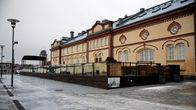En gul byggnad med stora fönster och tegeldetaljer står vid en öppen stenbelagd yta.