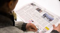 View over the shoulder of a person reading a tabloid sized newspaper on a white table.