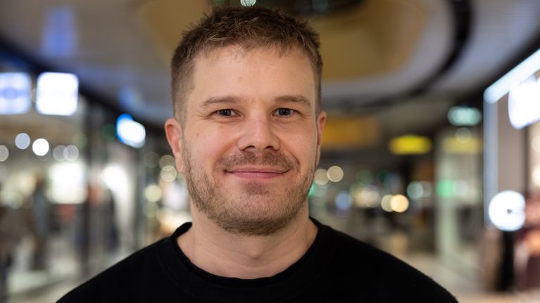 Mustaan paitaan pukeutunut mies seisoo kauppakeskuksen käytävällä ja katsoo kameraan hymyillen.