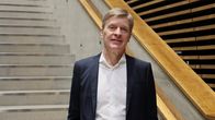 En person som står på en trappa i en modern byggnad med träpaneler. Han har på sig en mörk kostym och vit skjorta.