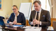 En man och en kvinna med sakliga kläder och bistra miner sitter vid ett långt mötesbord.