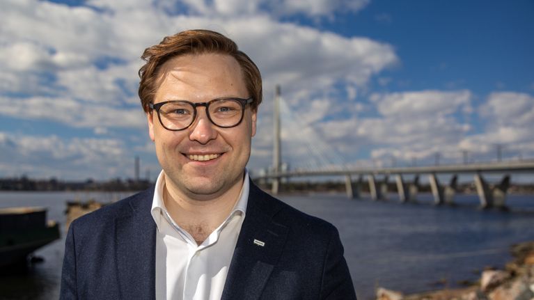 En person i kostym står utomhus vid vatten med en bro i bakgrunden.