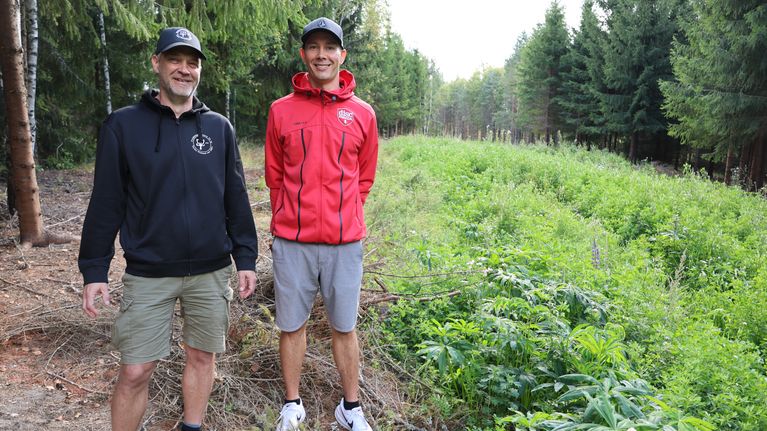 Två personer står på en stig i skogsmiljö, omgivna av träd och grön vegetation.