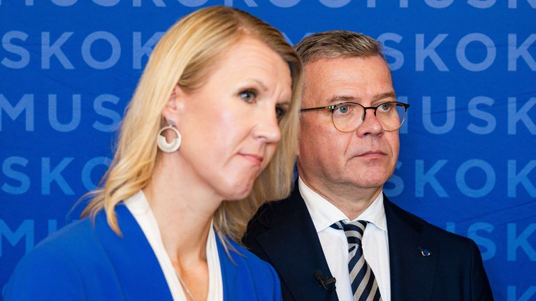 Kaksi ihmistä seisoo sinisen taustan edessä, jossa toistuu sana ”KOKOOMUS”.