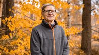 Harmaaseen takkiin pukeutunut Hannu Välimaa seisoo syksynsävyisten puiden edustalla ja hymyilee kameralle.