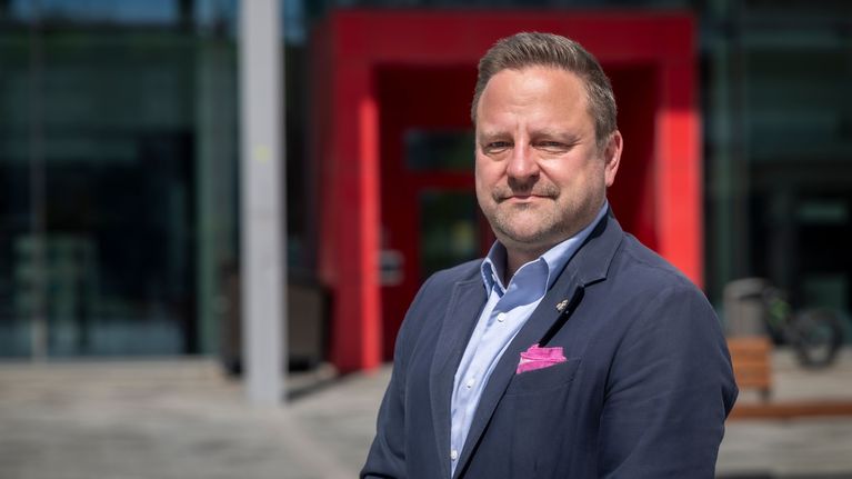 Mikko Aaltonen, en person i kostym står utomhus framför en byggnad med en röd entré.