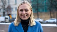 Siniseen takkiin pukeutunut Elina Valtonen seisoo aukiolla ja katsoo kameraan hymyillen.