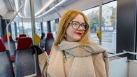 Nainen seisoo raitiovaunun sisällä pitämässä kiinni tangosta, ympärillä tyhjiä istuimia, taustalla kaupunkimaisema.