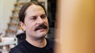 En man med mustasch och uppsatt hår står inomhus framför en suddig, ljus yta.