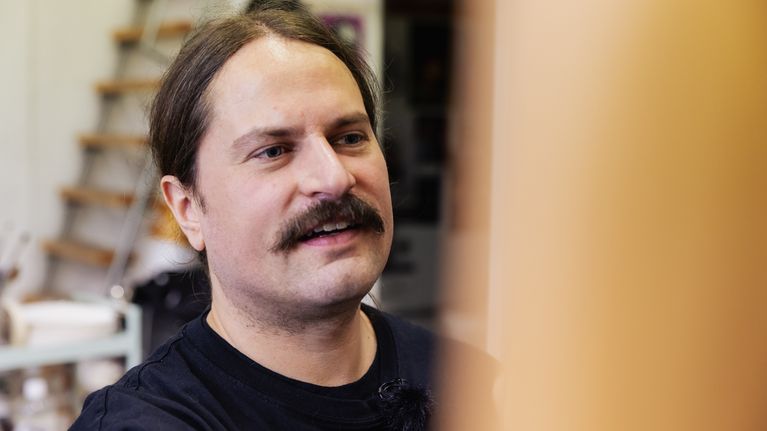 En man med mustasch och uppsatt hår står inomhus framför en suddig, ljus yta.