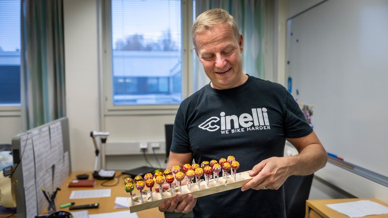 Antti Kontula seisoo työhuoneessaa ja hänellä on kädessään lauta, johon on kiinnitetty tikkukaramellejä.