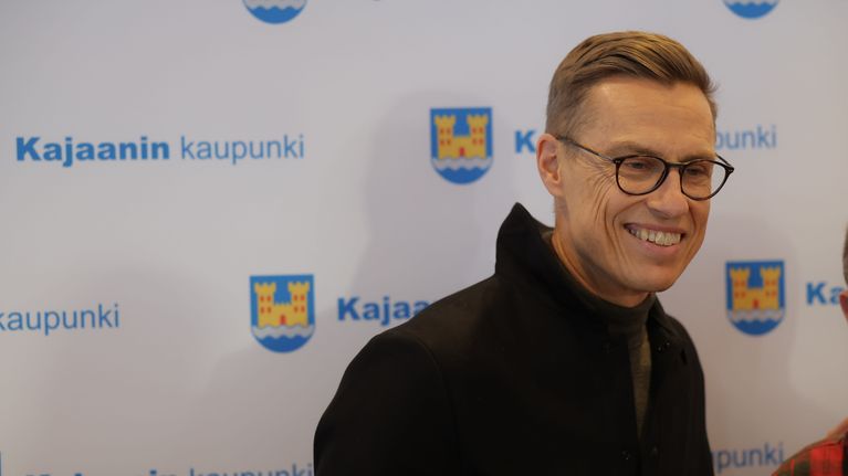 Tasavallan presidentti Alexander Stubb hymyilee, taustalla lakana, jossa on Kajaanin kaupungin logoja.