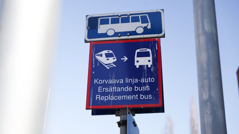 Tolpassa ylinnä sinivalkoinen liikennemerkki bussipysäkistä, lisäkilpenä korvaava linja-auto -teksti suomeksi, ruotsiksi ja englanniksi.