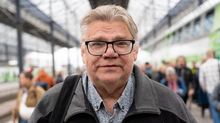 Timo Soini står på järnvägsstation och tittar i kameran. 