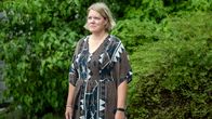 En kvinna med ljust axellångt hår står utomhus framför gröna buskar, iklädd mönstrad klänning.